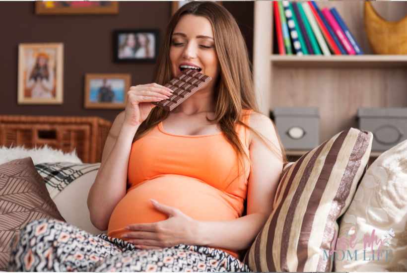 Pregnant woman in orange shirt relaxing on couch eating a chocolate bar